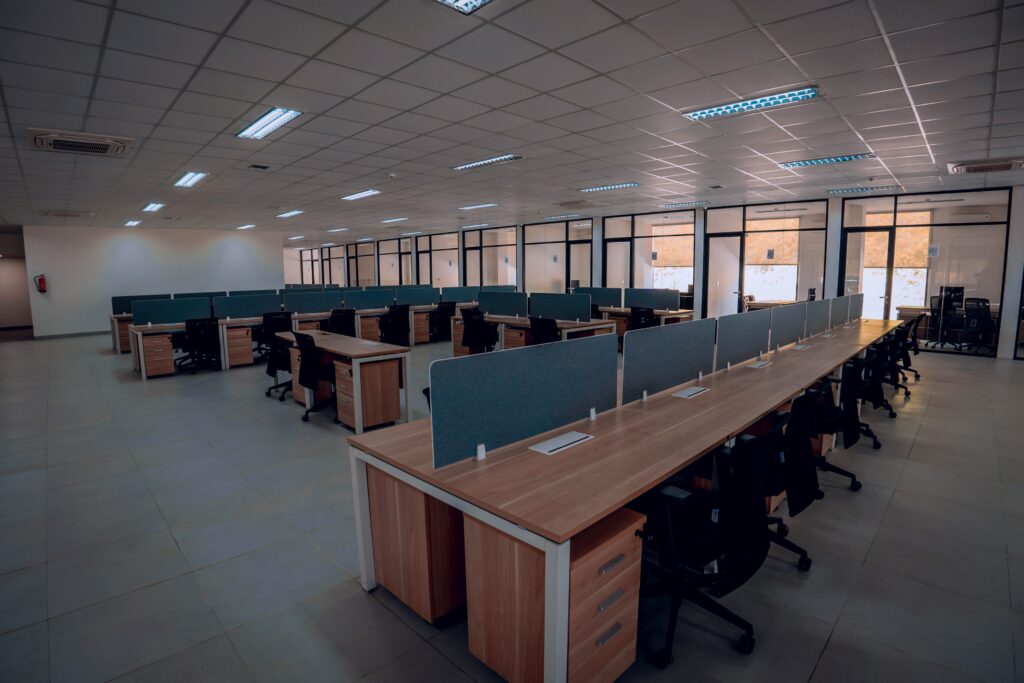 spacious modern office interior featuring rows of wooden desks with partition panels, ergonomic black chairs, and large glass-walled meeting rooms in the background under bright ceiling lights