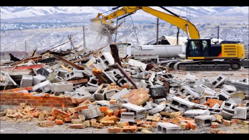 Excavator clearing construction debris and rubble from a demolished building site with piles of bricks and concrete blocks.