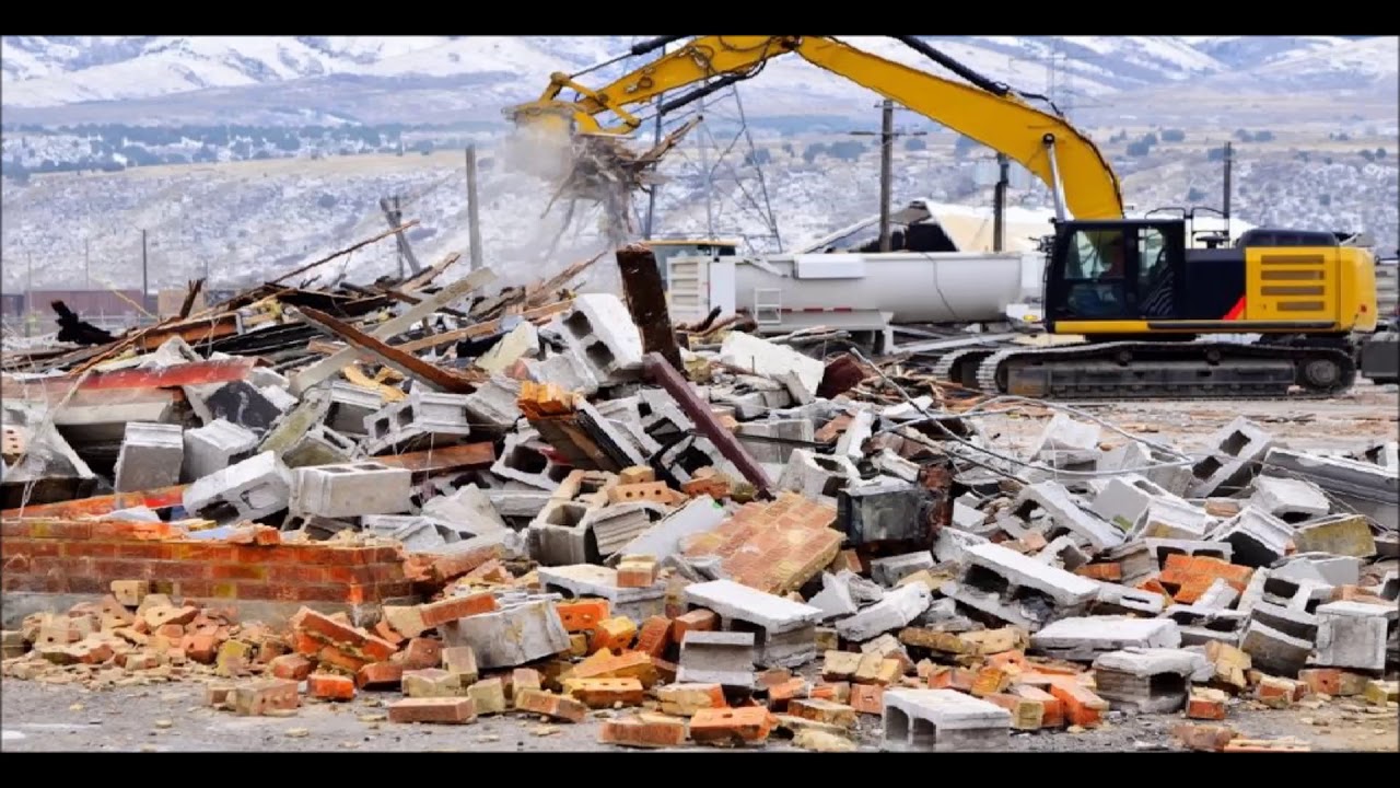 Excavator clearing construction debris and rubble from a demolished building site with piles of bricks and concrete blocks.